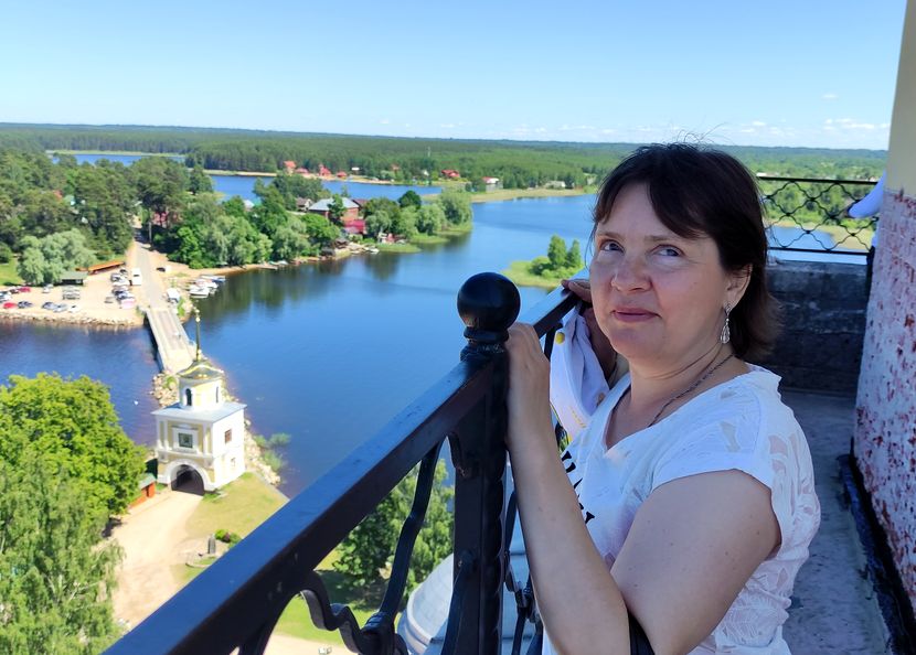 женщина в светлой футболке стоит на балконе высокого здания, держась рукой за кованые перила. перед ней внизу река, небольшая часовня, мост через реку
