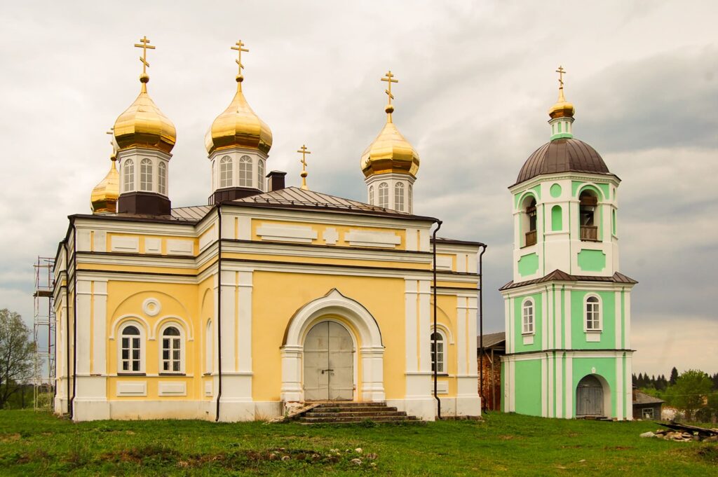 здание мужского монастыря нило-столобенской пустыни. желто-белое одноэтажное здание с золотыми куполами и крестами. справа зеленая, двухэтажная узкая колокольня
