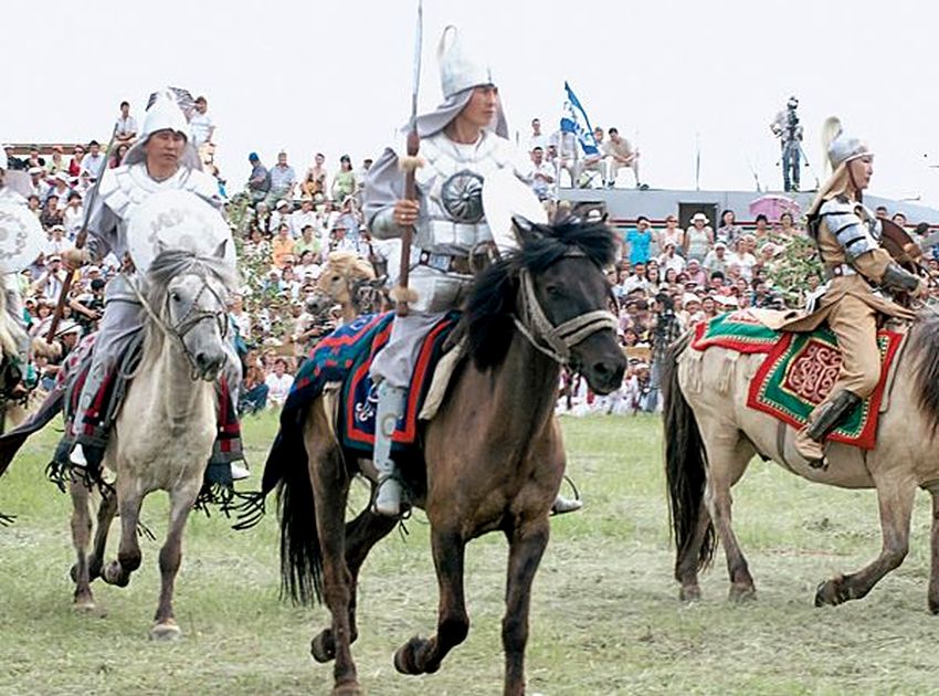 три всадника на конях скачут по стадиону в национальных одеждах, похожих на серебристые доспехи и остроконечные шлемы, в руках у двоих копья. позади них много зрителей на трибунах