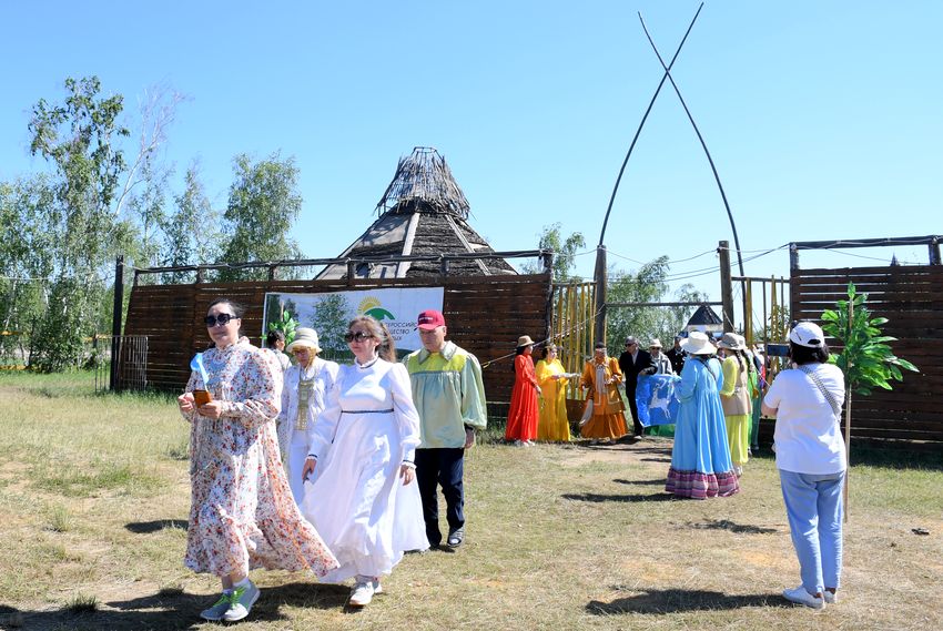 деревянный забор, на нем баннер с логотипом и надписью вос. из ворот выходит процессия в национальных костюмах, это члены якутской ро вос