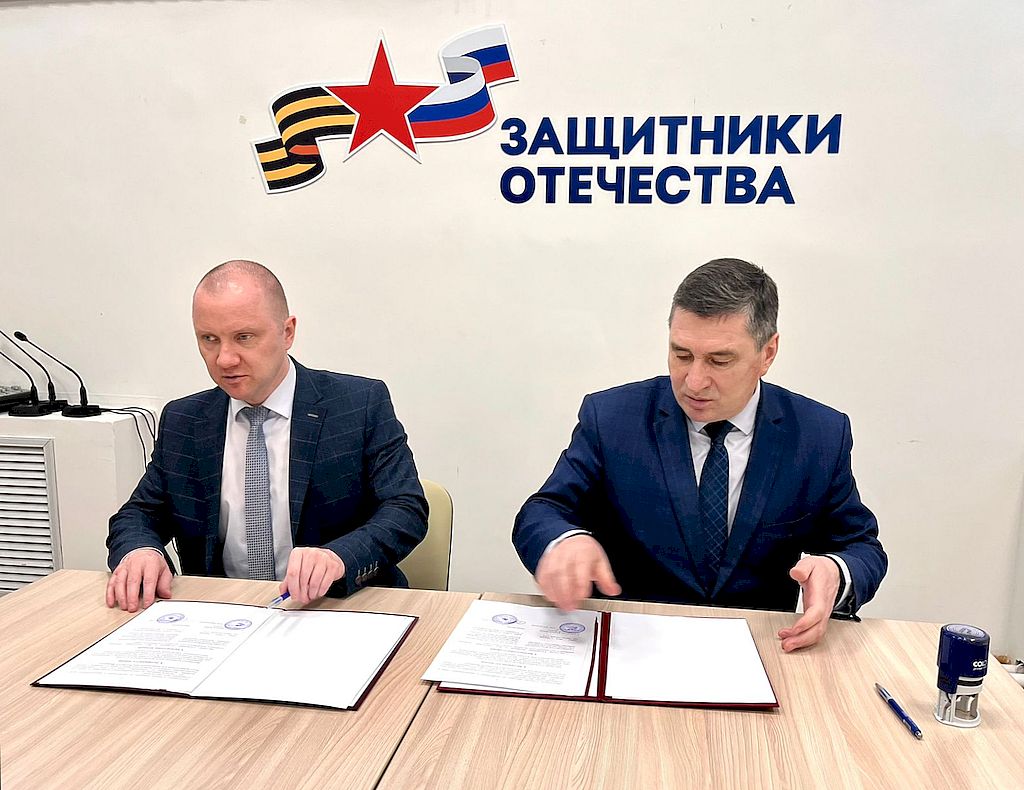 О. В. Шигирданов и А. М. Орисенко сидят за столом. Перед ними документы, в которых они ставят свои подписи.