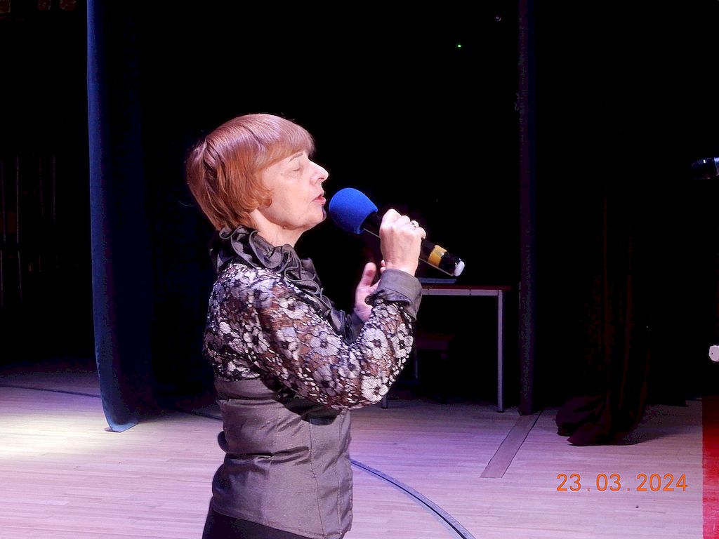 Участница регионального этапа на сцене. Фотография сделана сбоку. На женщине тёмная кофта с кружевным воротником-стойкой. В руках - микрофон. 