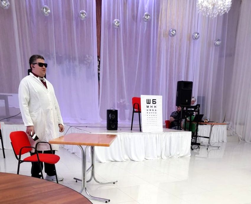 молодой мужчина в черных очках и белом медицинском халате. перед ним стул и  стол, справа небольшая сцена, на которой стоит прислоненная к стулу таблица для проверки остроты зрения