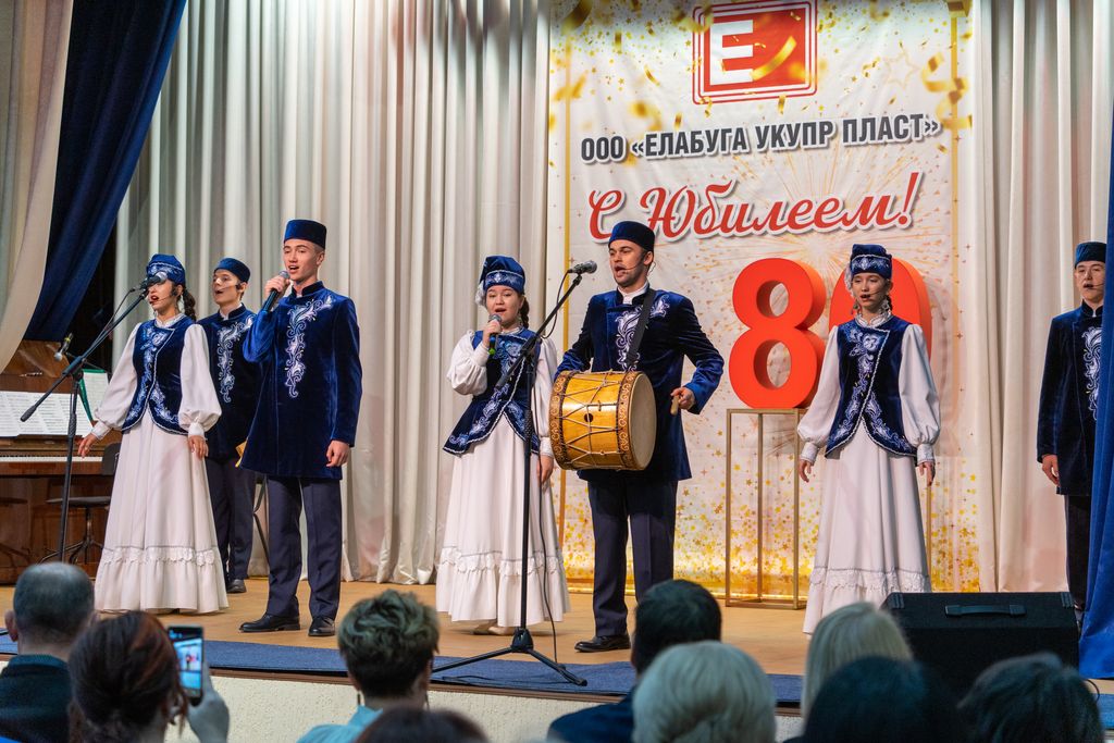 На сцене коллектив художественной самодеятельности - молодые люди в татарских национальных костюмах бело-синего цвета. На заднем плане баннер с надписью: "ООО Елабуга Укупрпласт" С юбилеем!" Перед баннером  на подставке объёмное красное число "80" .