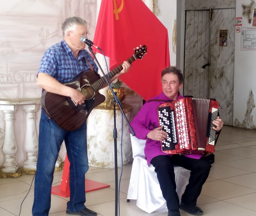 Слева мужчина стоя играет на гитаре и поёт в микрофон, справа мужчина, сидя играет на баяне. За ними висит красный флаг с серпом и молотом в верхнем левом углу полотнища.