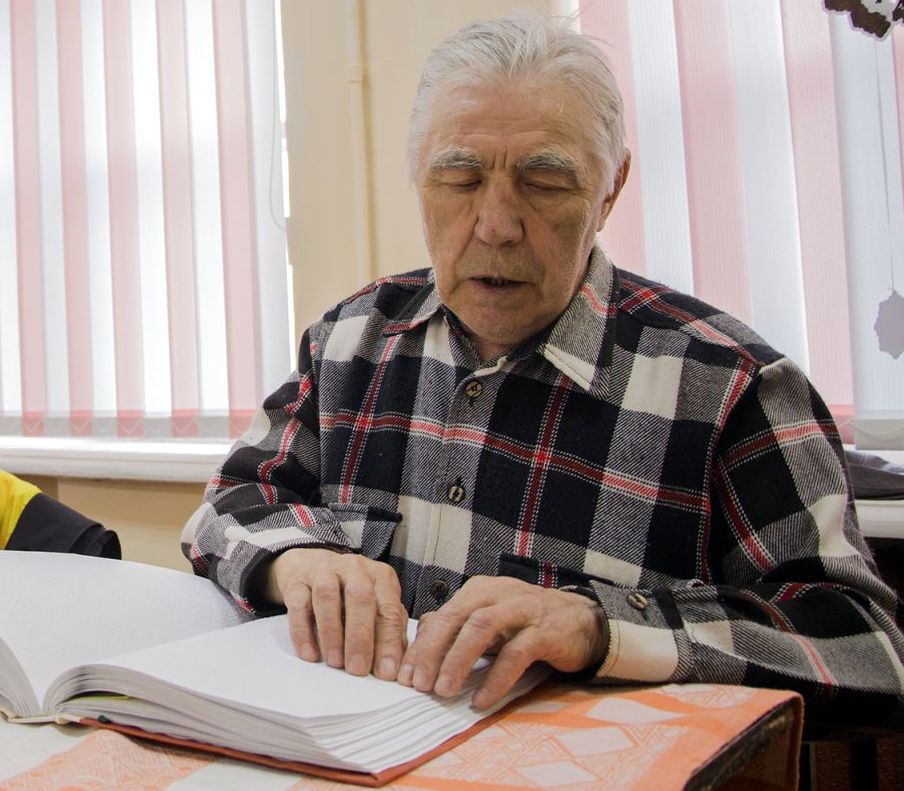 Участник конкурса - пожилой седой мужчина в клетчатой рубашке сидит за столом, руки на раскрытой книге, написанной шрифтом Брайля, глаза закрыты.