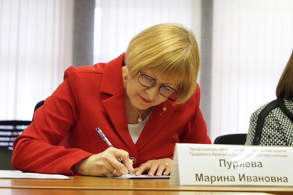 М. И. Пуряева - женщина в очках со светлыми короткими волосами, стрижка каре, в ярко-красном костюме. Подписывает документ, сидя за столом. Перед ней именная табличка.