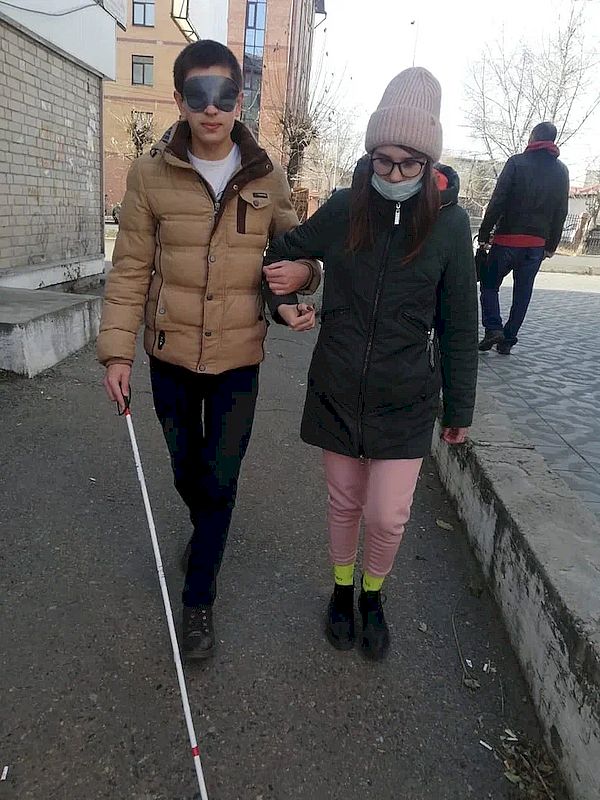 молодой человек с повязкой и тростью идёт по дороге. Под руку его держит девушка.