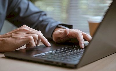 на фотографии открытый ноутбук, за которым сидит человек. Его руки лежат на клавиатуре