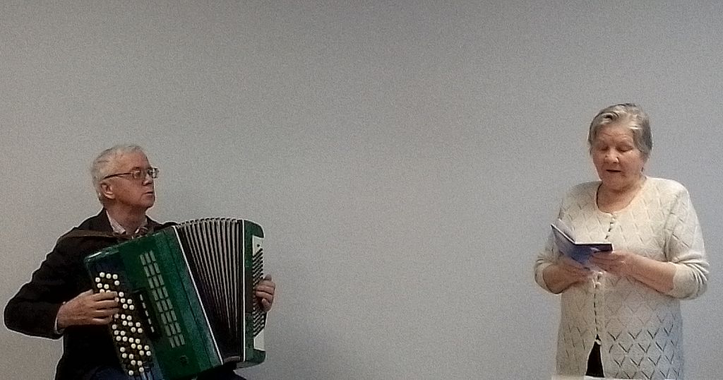 на фотографии двое. Женщина читает стихи, мужчина играет на баяне