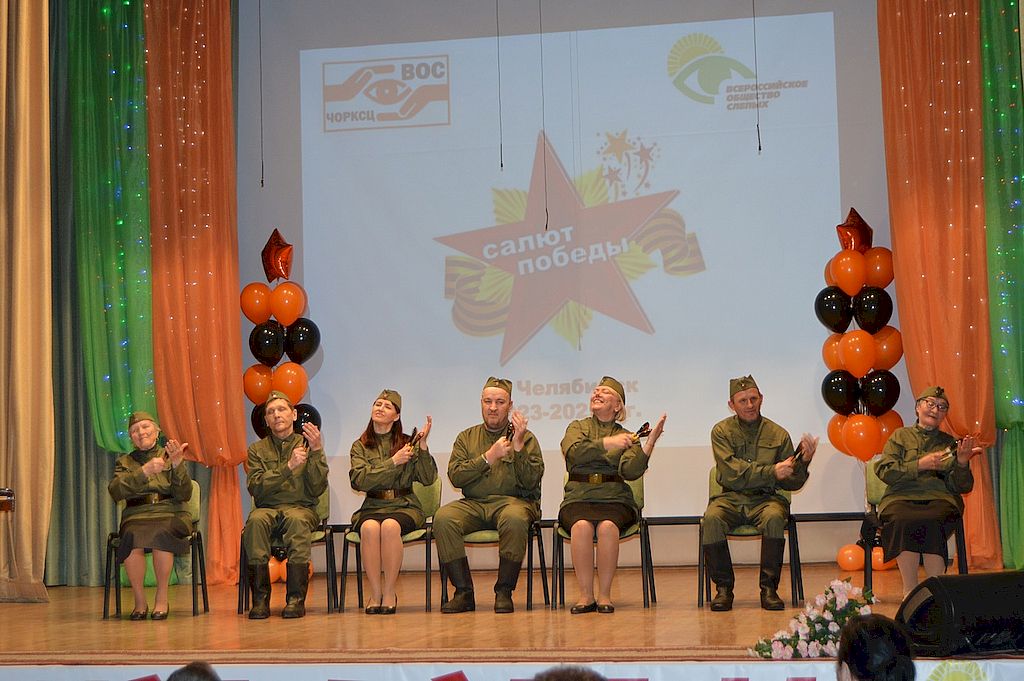 Участники фестиваля сидят на сцене и играют на ложках. Они одеты в гимнастёрки. 