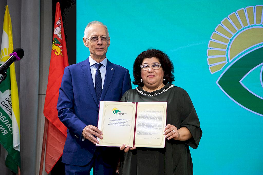 Вице-президент ВОС Александр Коняев на сцене актового зала санатория ВОС «Сосны» передает и.о. председателя МОО ОООИ ВОС Мтварисе Датикашвили поздравительный адрес от президента ВОС Владимира Сипкина. На стене установлены флаги Московской области и Московской областной организации ВОС