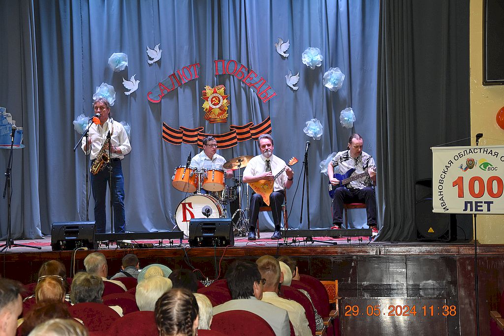 На сцене квартет из мужчин - саксофон, гитара, барабаны и балалайка. На заднике сцены надпись "Салют Победы", белые объёмные цветы и птицы. 
