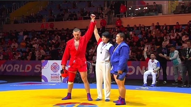 Чемпион мира с поднятой рукой вверх в красном костюме. Рядом с ним судья и спортсмен, занявший второе место