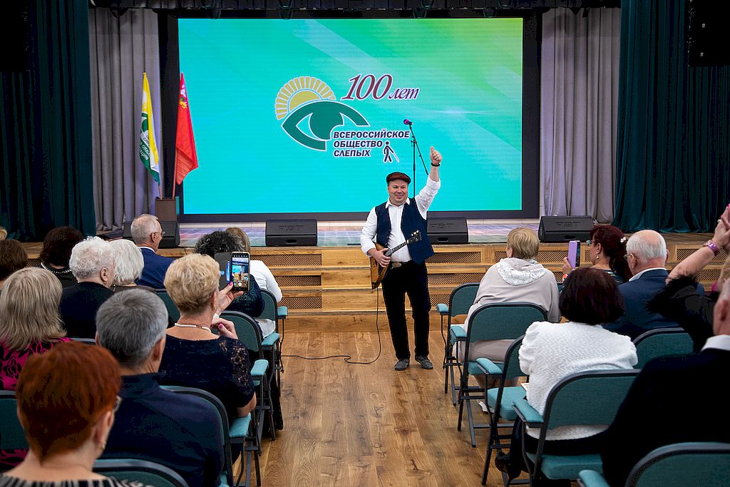 Актовый зал санатория ВОС «Сосны». У первых рядов зрительного зала виртуоз-балалаечник жестом «большой палец вверх» подтверждает высокий уровень проведения юбилейного мероприятия в честь 100-летия ВОС и Московской областной организации ВОС