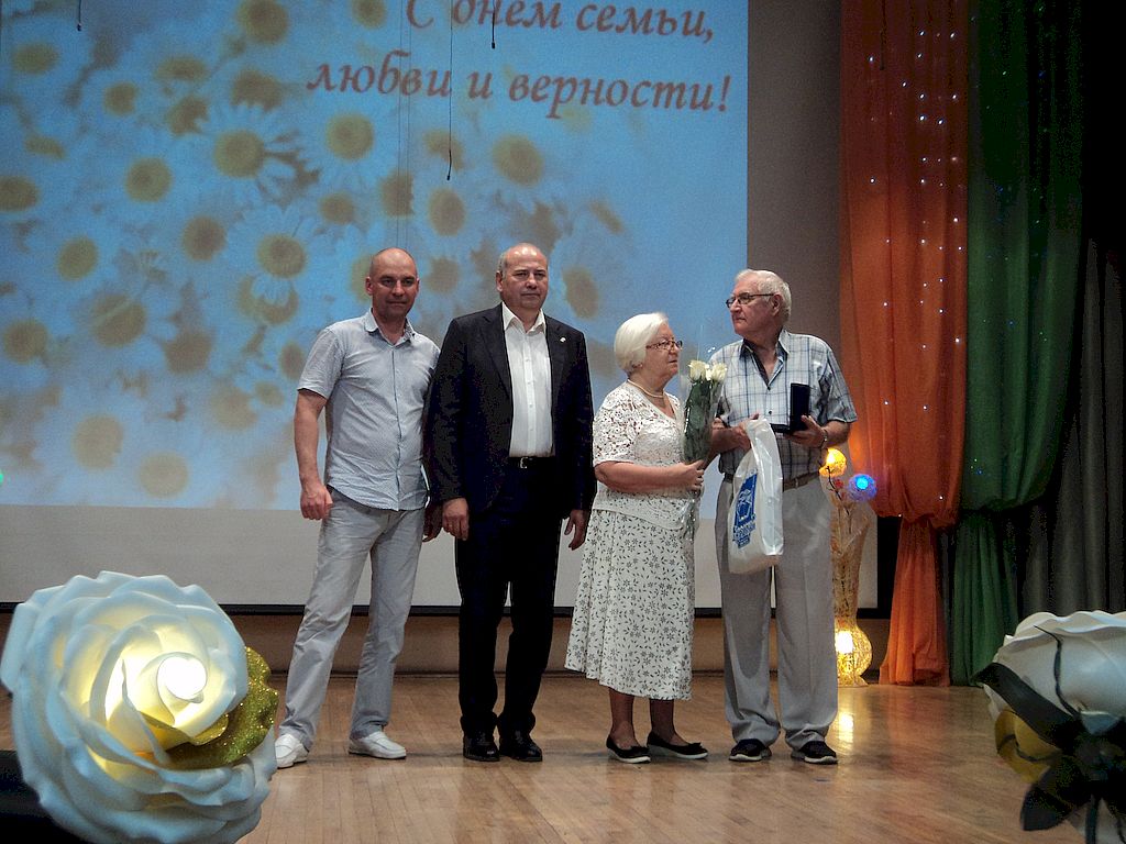 На сцене семейная пара. У них в руках цветы, пакет с подарками и коробочка с медалями.  Рядом стоят двое мужчин.