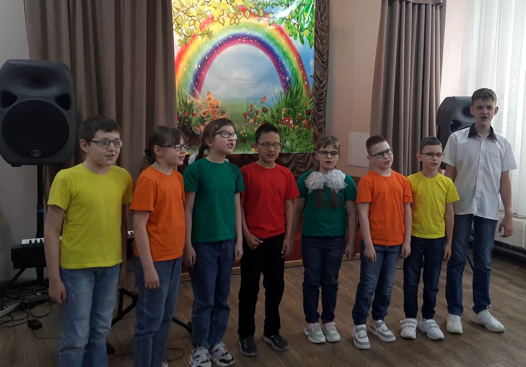 Мальчики и девочки исполняют песняю, всего 8 человек. На них джинсы, кроссовки и разноцветные футболки. Семь из восьми человек в очках. На заднем плане: звуковые колонки, кулисы и баннер с изображением деревьев, радуги, цветов.