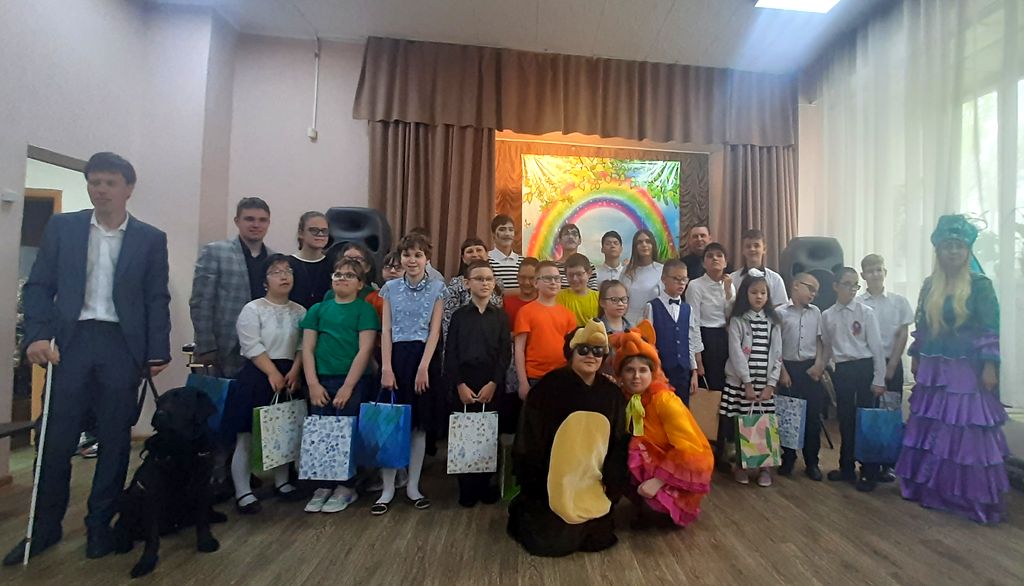 Групповое фото участников фестиваля: мальчики и девочки разных лет, с подарочными пакетами в руках. Слева в первом ряду стоит молодой человек с белой тростью и чёрным лабрадором на шлейке. На переднем плане две девушки в костюме медведя и лисы.