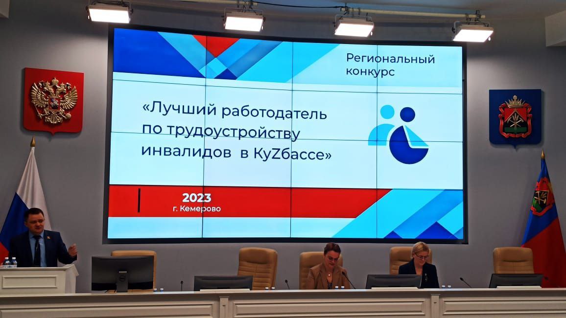 Большой плазменный экран с надписью: "Региональный конкурс. Лучший работодатель по трудоустройству инвалидов в КуZбассе. Кемерово, 2023". Перед экраном трибуна и стол для заседания. Слева за трибуной с микрофоном стоит молодой мужчина в костюме и галстуке. За ним стоит флаги герб РФ. По центру перед экраном за столом сидят две женщины, перед ними мониторы и микрофоны для конференции. Справа от них герб и флаг Кемеровской области.