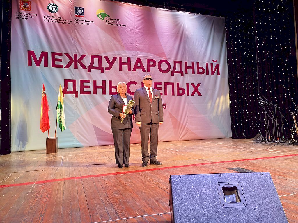 Сцена Делового и культурного центра «Костино». В центре сцены – член Центрального Правления ВОС, депутат Московской областной Думы, заместитель председателя Комитета по социальной политике и здравоохранению Владимир Вшивцев в деловом костюме серого цвета с орденскими планками и депутатским нагрудным знаком. Слева его помощник – член Бюро Химкинской местной организации ВОС Ирина Палёнова после торжественного вручения ей Юбилейной медали «100 лет образования Всероссийского общества слепых». Ирина Палёнова держит в руках Юбилейную медаль и букет цветов.  На заднем плане во всю стену растяжка с надписью в центре «Международный день слепых». Вверху растяжка с изображением (слева направо) Герба Московской области, эмблемы Минсоцразвития Московской области, Герба города Королёв и эмблемы Всероссийского общества слепых. На сцене слева на флагштоках установлены флаги Московской области и Московской областной организации ВОС.  