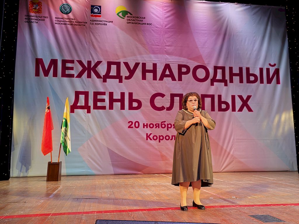 На сцене с заключительным словом выступает и.о. председателя МОО ОООИ ВОС Мтвариса Датикашвили в элегантном платье коричневого цвета. В руках – микрофон. На заднем плане во всю стену растяжка с надписью в центре «Международный день слепых». Вверху растяжка с изображением (слева направо) Герба Московской области, эмблемы Минсоцразвития Московской области, Герба города Королёв и эмблемы Всероссийского общества слепых. На сцене слева на флагштоках установлены флаги Московской области и Московской областной организации ВОС. 