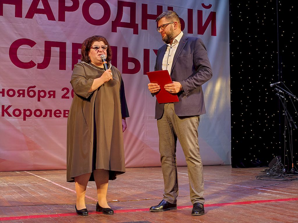 На сцене выступает и.о.председателя МОО ОООИ ВОС Мтвариса Датикашвили в элегантном платье коричневого цвета. В ее руках – микрофон. Слева председатель Московской областной региональной организации Всероссийского общества глухих Дмитрий Жевжиков в деловом костюме. Он держит в руках папку алого цвета с поздравительным адресом.