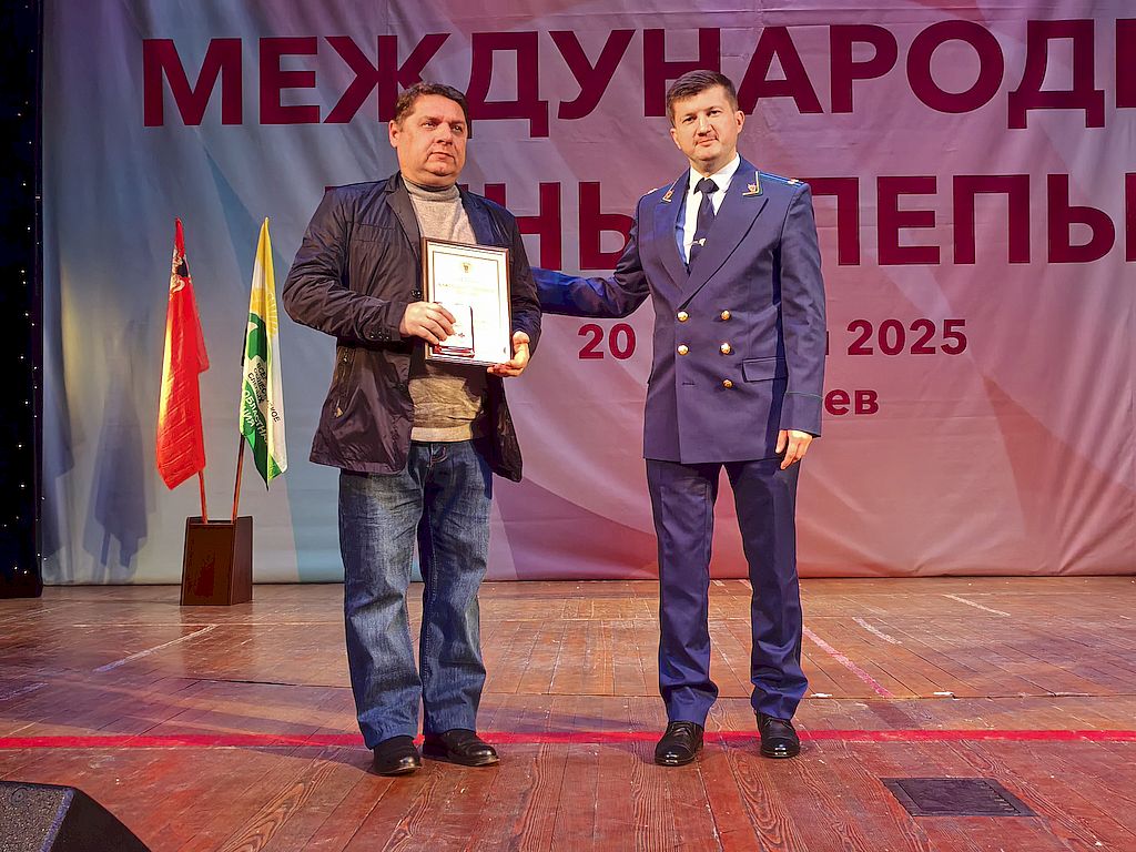 На сцене – прокурор отдела по надзору за соблюдением федерального законодательства, прав и свобод гражданина Управления по надзору за исполнением федерального законодательства, советник юстиции Сергей Гончаров в прокурорской форме. Он вручает Благодарственное письмо прокурора Московской области секретарю Подольской местной организации ВОС Вячеславу Чернецкому (слева). Награждаемый в джинсах, бежевом свитере и темно-серой куртке. Он держит в руках Благодарственное письмо и Юбилейную медаль ВОС. 