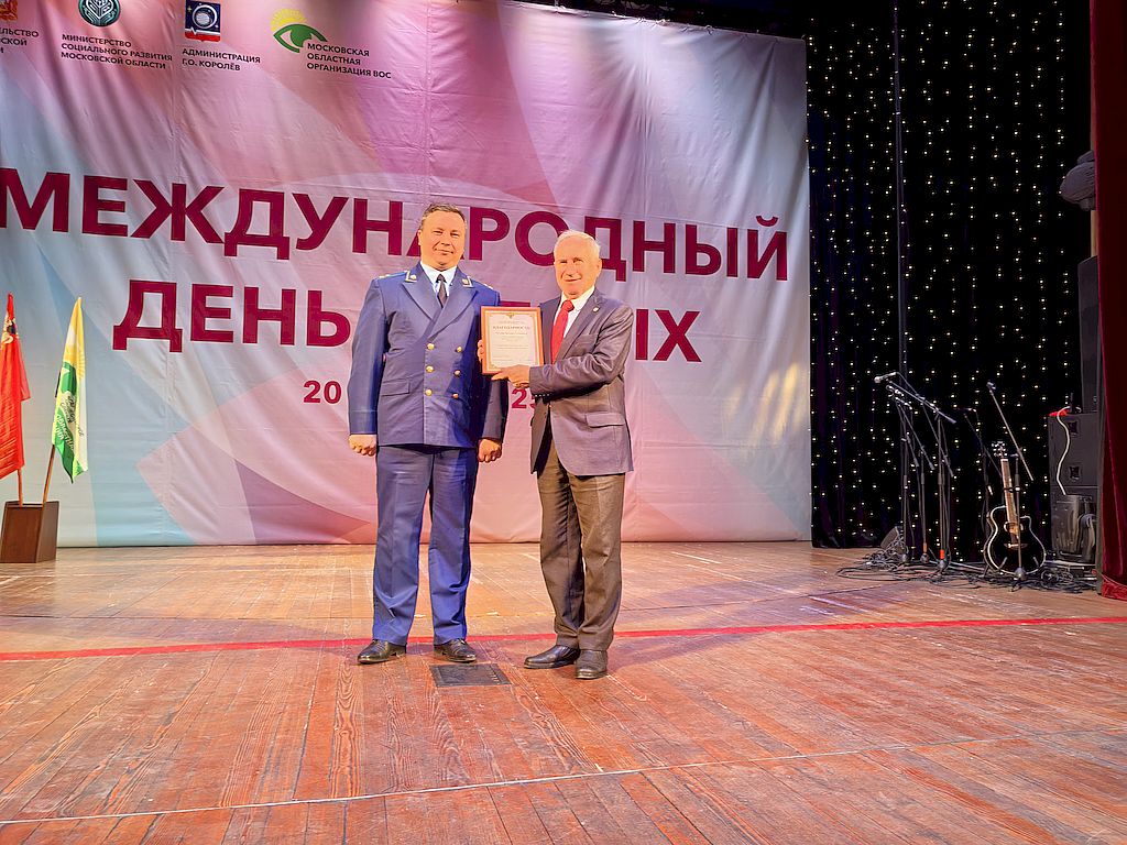 На сцене – Московско-Смоленский транспортный прокурор, старший советник юстиции Сергей Портянов в прокурорской форме. Он вручает ведущему специалисту Аппарата управления МОО ОООИ ВОС Валерию Рягузову (справа) Благодарность Московско-Смоленской транспортной прокуратуры. Валерий Рягузов в серых брюках и классическом пиджаке темно-синего цвета. На заднем плане на флагштоках флаги Московской области и Московской областной организации ВОС