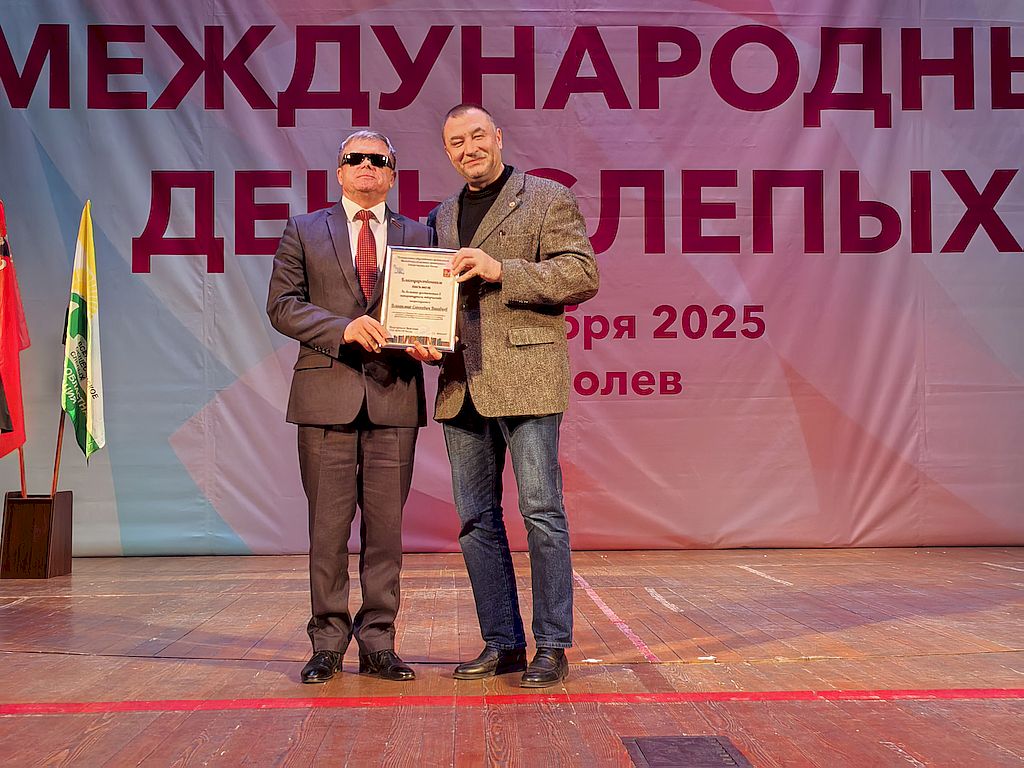 На сцене председатель Московского областного отделения Союза писателей России Сергей Антипов в брюках джинсового типа, черной футболке и свободного кроя пиджаке двух оттенков серого с нагрудным знаком Союза писателей. Он вручает Благодарственное письмо Отделения Союза писателей России члену ЦП ВОС, депутат Московской областной Думы, заместителю председателя Комитета по социальной политике и здравоохранению Владимиру Вшивцеву (слева). Владимир Вшивцев в деловом костюме серого цвета с орденскими планками и депутатским нагрудным знаком. На заднем плане на флагштоках флаги Московской области и Московской областной организации ВОС