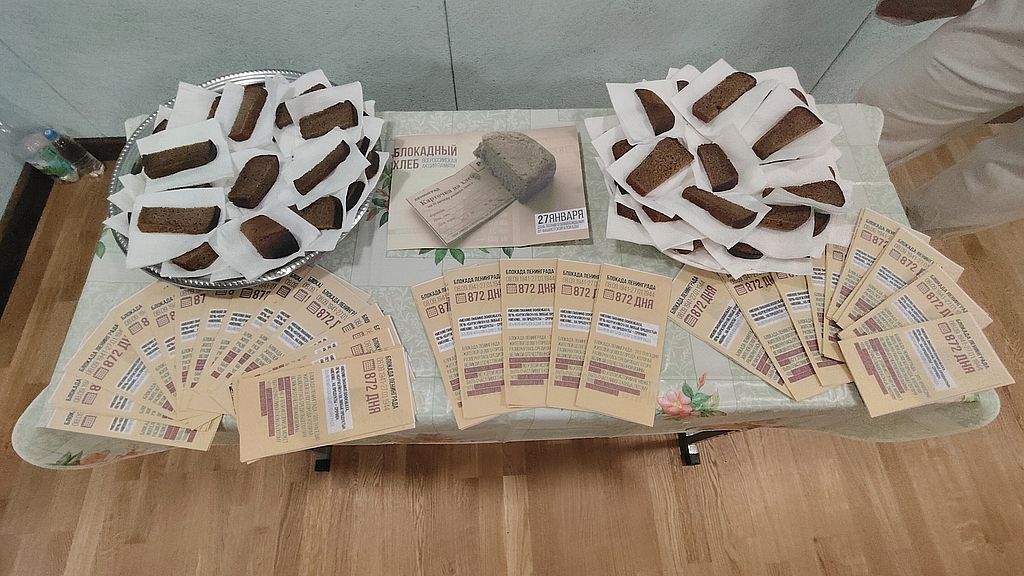 на столе лежит изображение блокадного хлеба. Рядос в тарелках лежат куски чёрного хлеба