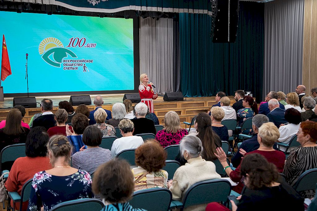 Артистка в русском народном платье исполняет патриотичную песню в актовом зале санатория ВОС «Сосны». На сцене расположен экран с логотипом столетия ВОС