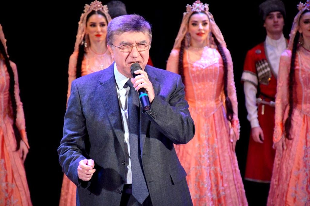 Д. М. Магомедов на сцене с микрофоном, за ним стоят девушки в национальных костюмах.