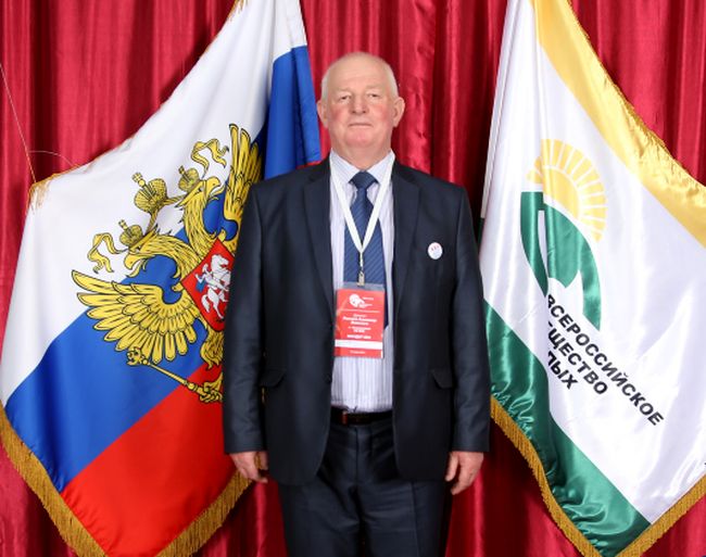 Цветная портретная фотография А. Я. Романова. На красном фоне висят флаги РФ и ВОС. Посередине стоит взрослый мужчина в костюме и галстуке. На груди висит бейдж.