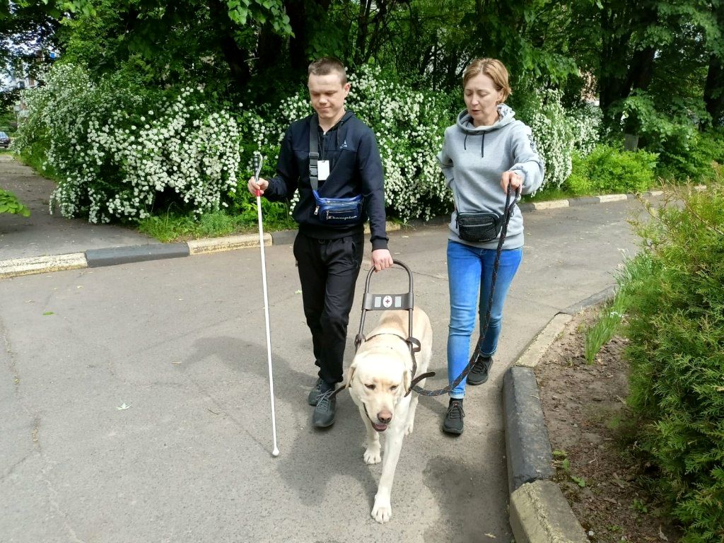 Молодой мужчина с закрытыми глазами с белой тростью идёт по парку с собакой-проводником. Справа от него женщина инструктор.