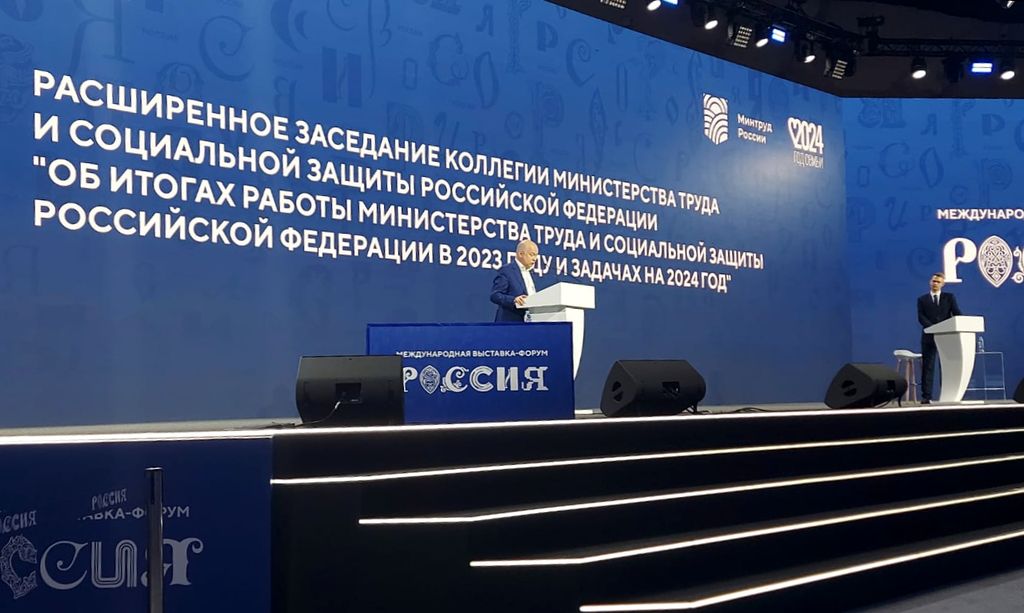 Мужчина в костюме стоит возле белой трибуны и читает доклад. На заднем плане на синем фоне надпись: "Расширенное заседание коллегии Министерства труда и соцзащиты по теме «Итоги работы Министерства труда и социальной защиты Российской Федерации в 2023 году и задачах на 2024 год».