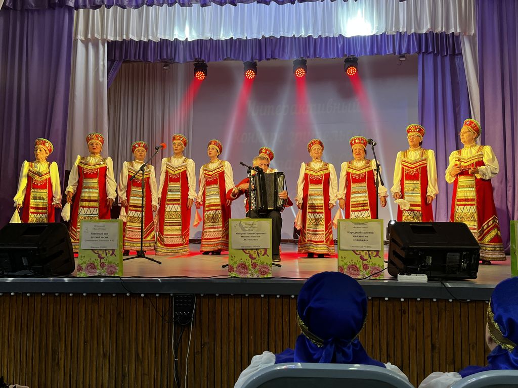 Выступление народного хора русской песни. Десять взрослых женщин в народных красных сарафанах и кокошниках стоят на сцене перед микрофонами. Мужчина в красной рубашке, сидя, играет на баяне.