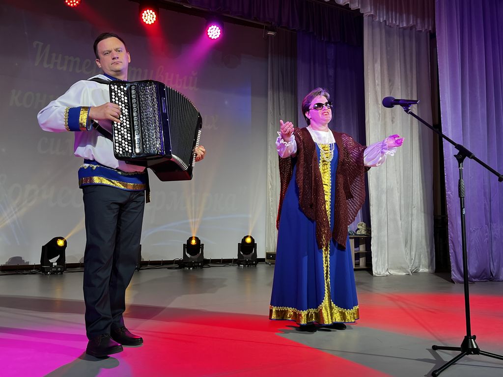 мужчина в русской белой с синей отделкой рубашке играет на баяне, рядом справа женщина в темных очках, в синем русском сарафане, раскинув в стороны руки поет у микрофона. 
